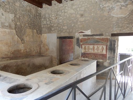 Casa e Thermopolium di Vetutius Placidus.  The complex (as its name indicates) is consisted of a private house at the back and a bar/restaurant located on the road, where drinks and hot food were served. In the shop one can see the large jars placed in the richly decorated masonry counter. The lalarium (a painted shrine resembling a temple) on the back wall is well-maintained, and represents the Lares, as well as god Mercury and god Dionysus.
