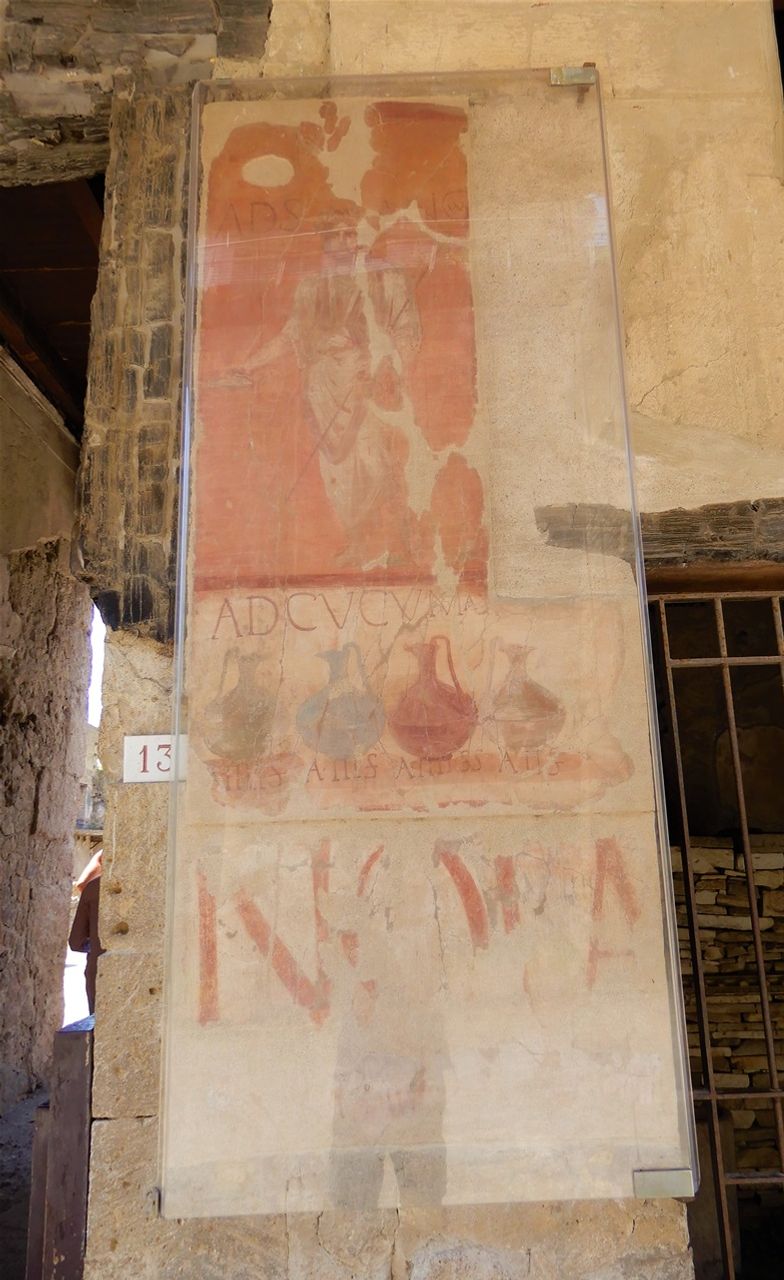 Cucumas's shop (Bottega ad Cucumas). Cucumas's shop, or inn, bears a wall painting by its entrance which announces the prices of pitchers of wine, along with illustrations. At the top can be seen the figure of Semo Sanctus, a divinity often compared to Hercules and who was said to protect business, with the inscription “ad Sancum”.  The panel at the bottom with the inscription NOLA in red letters is the announcement of a show.