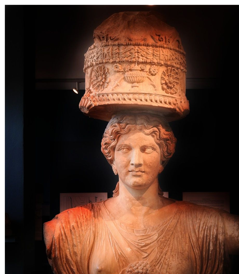 The upper part of one of the caryatids (Kore) that flanked the Lesser Propylaea of the sanctuary of Demeter and Kore at Eleusis. The caryatid was made in Attica in about 50 BC. (Eleusis Museum). Kore  brings on her head the ciste, the container holding the sacred articles of the ceremony, with a relief appearance of the symbols of the Eleusinian cult, which are: the ear of grain, the poppies, the rozetes and the kernos.
