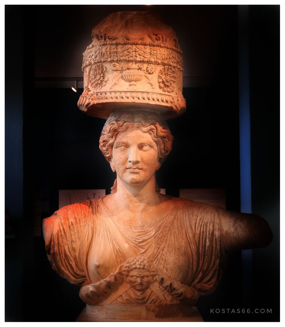 The upper part of one of the caryatids (Kore) that flanked the Lesser Propylaea of the sanctuary of Demeter and Kore at Eleusis. The caryatid was made in Attica in about 50 BC. (Eleusis Museum). Kore  brings on her head the ciste, the container holding the sacred articles of the ceremony, with a relief appearance of the symbols of the Eleusinian cult, which are: the ear of grain, the poppies, the rozetes and the kernos.